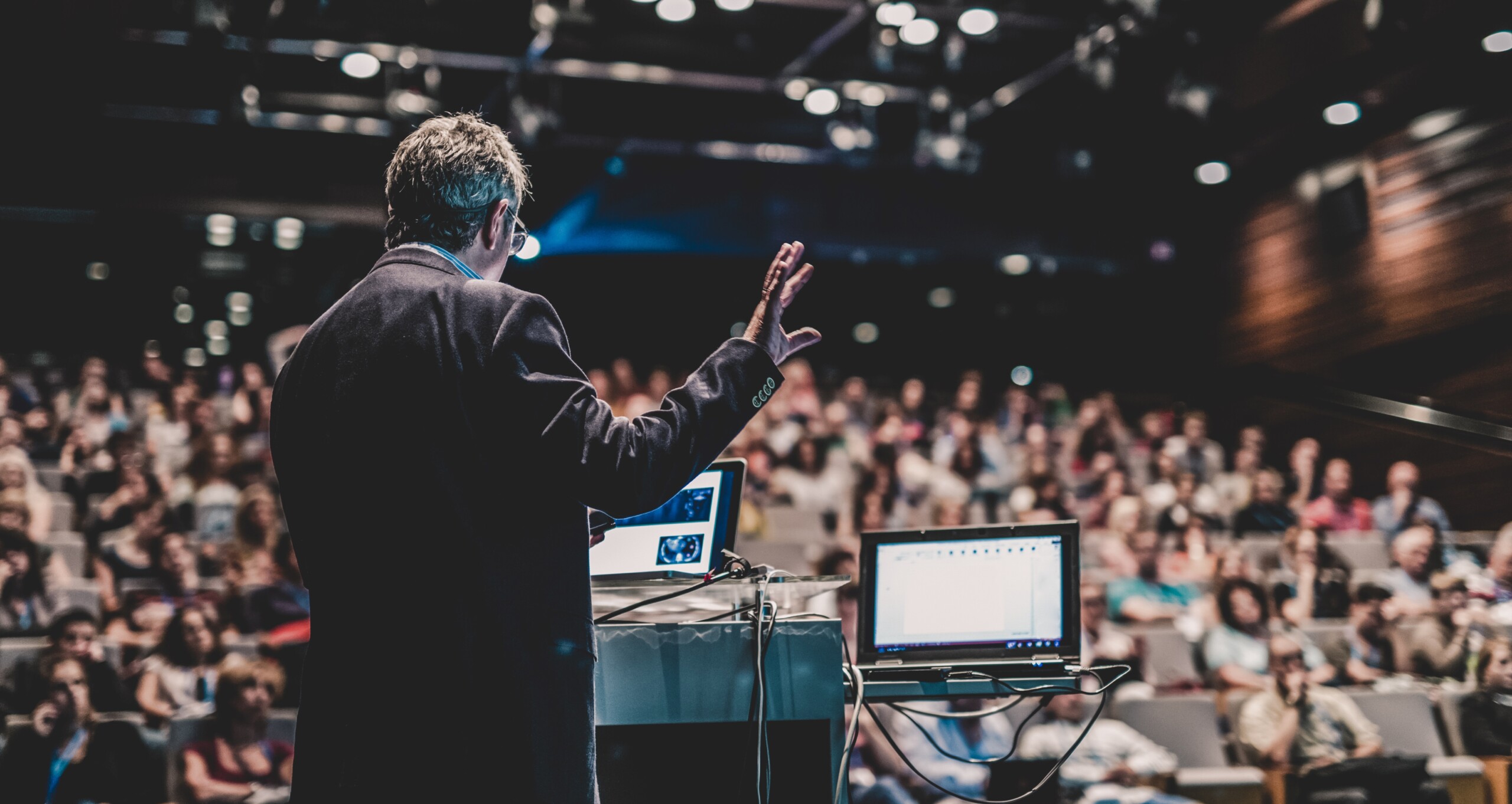 A person presenting a hybrid event, with a room full of people, and a couple of screens.